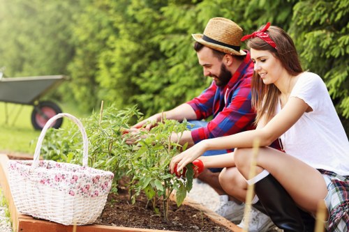 In a London garden, a young man and woman are engaged in gardening activity, kneeling beside a raised flower bed. The man, wearing a straw hat and a red and blue checkered shirt, is using his hands to tend to green plants. The woman, dressed in a white t-shirt and plaid skirt with a red headband, is also planting or inspecting the greenery. In the foreground, a white wicker basket filled with pink flowers sits on the edge of the flower bed. The garden features a neat, well-maintained lawn, with a background of lush green trees and shrubs, creating a peaceful outdoor scene typical of Fitzrovia. The lighting suggests a bright, sunny day, ideal for outdoor maintenance and gardening work associated with professional gardening services such as Lawn Mowing Fitzrovia, serving residential outdoor spaces in the local area.