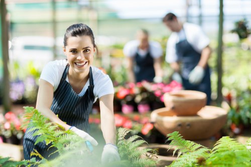A young woman with dark hair tied back, wearing a striped apron and white gloves, is smiling at the camera while tending to plants in a greenhouse or nursery setting. She is surrounded by vibrant green foliage, including ferns and other leafy plants, with soft natural light illuminating the scene. In the background, two other gardeners or staff members are visible, wearing similar aprons and working amongst colorful flower pots and containers. The environment suggests a professional gardening or landscaping setting, with organized plant displays, stone or terracotta water features, and a structured layout of garden beds and walkways, indicative of services offered by Lawn Mowing Fitzrovia in the London area, possibly within a garden centre or nursery designed for outdoor plant care, landscaping, and garden maintenance.