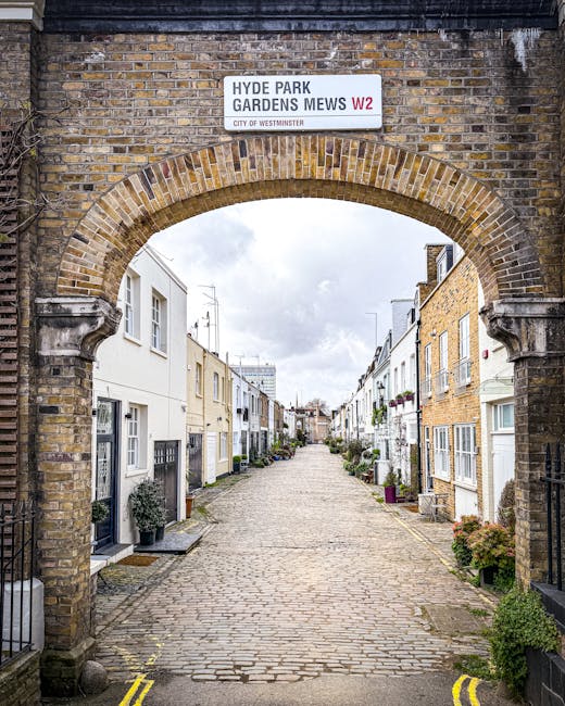 A cobbled alleyway in Fitzrovia mews gardens viewed through a brick archway, with a white sign reading 'Hyde Park Gardens Mews W2' positioned above the arch. The alley is lined with terraced houses pa