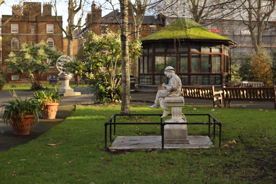 A peaceful garden scene featuring a neatly trimmed lawn bordered by paved walkways and lush green plants, including potted ferns and small shrubs, with a decorative stone statue of a young girl sittin